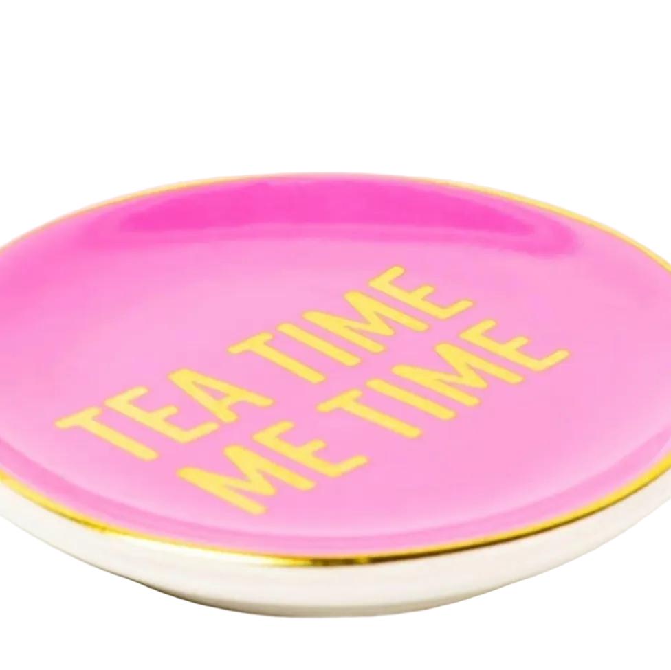 A round pink ceramic tea time keeper plate with gold lettering that reads 'TEA TIME ME TIME' and a gold rim, shown on a white background.